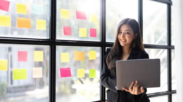 Asian freelance woman smiling paste post-it on the wall to record for work and using on laptop on wooden table at home. Entrepreneur woman working for her business at home. Business concept...