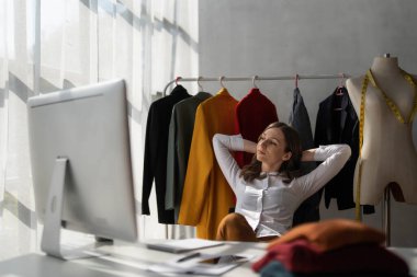 Full length portrait of attractive smiling young stylish fashion clothes designer sittng near table with computer, enjoying working in modern showroom atelier, casual workday concept...