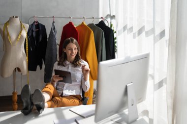 Full length portrait of attractive smiling young stylish fashion clothes designer sittng near table with computer, enjoying working in modern showroom atelier, casual workday concept...