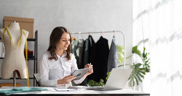 Дизайнер женской одежды, работающий с новой коллекцией женской одежды в уютной мастерской, модельер, портниха, стоящая рядом с вешалкой для одежды, трогательная, держащая в руках модную одежду ручной работы
