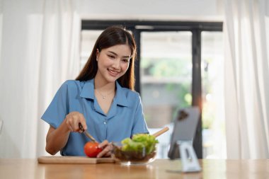 Asyalı güzel bir kadın mutfak arka planında otururken dizüstü bilgisayarla çalışıyor....