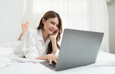 young asian woman using laptop when sitting on a bed. Smart living with communication technology for a better life. girl sitting on at white bedroom space with a clean interior design...