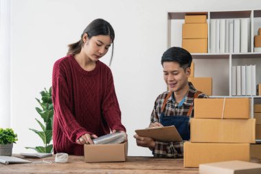 Genç Asyalı çiftler erkek ve kadın küçük işletme sahibi çevrimiçi siparişleri kontrol ediyor ve ürünü birlikte paketliyorlar.