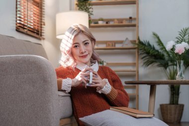 Genç ve güzel Asyalı kadın oturma odasında dinleniyor, kitap okuyor, kahve içiyor ve sabahları mutlu bir şekilde telefonu kullanıyor...