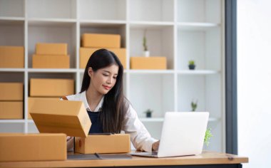 Genç bir Asyalı kadının portresi, arka planda paketlerle dolu bir ofiste oturan bir e-ticaret çalışanı KOBİ iş ekomünyesi ve teslimat işi için bir not ve hesap makinesi.