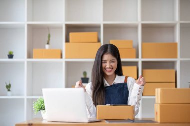 Genç bir Asyalı kadının portresi, arka planda paketlerle dolu bir ofiste oturan bir e-ticaret çalışanı KOBİ iş ekomünyesi ve teslimat işi için bir not ve hesap makinesi.