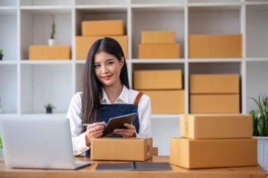Genç bir Asyalı kadının portresi, arka planda paketlerle dolu bir ofiste oturan bir e-ticaret çalışanı KOBİ iş ekomünyesi ve teslimat işi için bir not ve hesap makinesi.