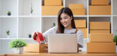 Genç bir Asyalı kadının portresi, arka planda paketlerle dolu bir ofiste oturan bir e-ticaret çalışanı KOBİ iş ekomünyesi ve teslimat işi için bir not ve hesap makinesi.