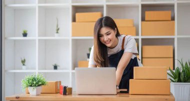 Genç bir Asyalı kadının portresi, arka planda paketlerle dolu bir ofiste oturan bir e-ticaret çalışanı KOBİ iş ekomünyesi ve teslimat işi için bir not ve hesap makinesi.