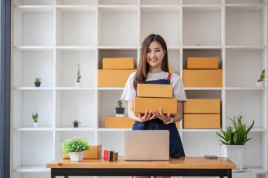 Genç bir Asyalı kadının portresi, arka planda paketlerle dolu bir ofiste oturan bir e-ticaret çalışanı KOBİ iş ekomünyesi ve teslimat işi için bir not ve hesap makinesi.
