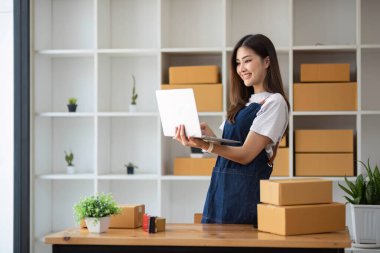 Genç bir Asyalı kadının portresi, arka planda paketlerle dolu bir ofiste oturan bir e-ticaret çalışanı KOBİ iş ekomünyesi ve teslimat işi için bir not ve hesap makinesi.