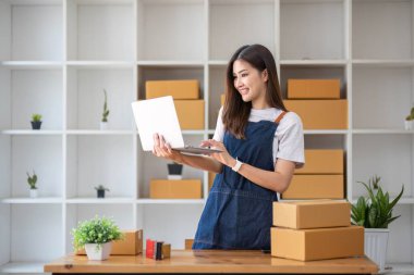 Genç bir Asyalı kadının portresi, arka planda paketlerle dolu bir ofiste oturan bir e-ticaret çalışanı KOBİ iş ekomünyesi ve teslimat işi için bir not ve hesap makinesi.