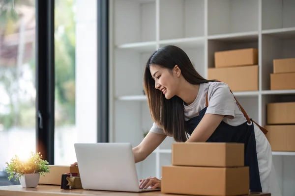 stock image A portrait of a young Asian woman, e-commerce employee sitting in the office full of packages in the background write note of orders and a calculator, for SME business ecommerce and delivery business