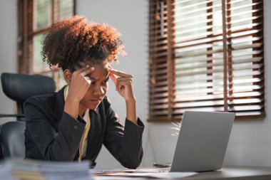 Afro saçlı genç Afrikalı bir kadın sinirli ve stresli görünüyor, masada oturuyor, dizüstü bilgisayar kullanıyor, düşünüyor ve kameraya bakıyor, depresyon sorunlarından yorgun ve sıkılmış hissediyor...