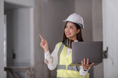 İnşaat alanında dizüstü bilgisayar tutan ve kaskını takan Mutlu profesyonel inşaat mühendisi kadın portresi, endüstriyel mühendislik konsepti..