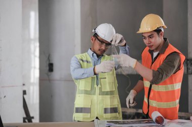 Yelekli ve şapkalı genç adam inşaat alanı inşaatı projesinde dizüstü bilgisayarla çalışıyor. Mühendis iş arkadaşlarıyla konuşuyor..