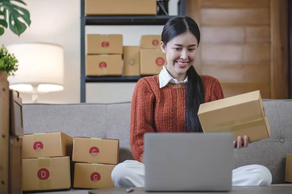 Asian business woman leaning on the sofa use laptop computer checking customer order online shipping boxes at home. Starting Small business entrepreneur SME. Online business, Work at home concept...