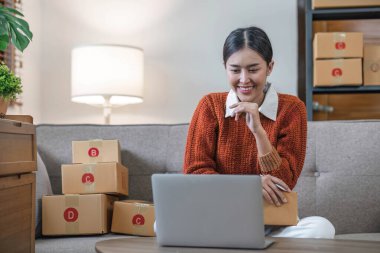 Kanepeye yaslanan Asyalı iş kadını dizüstü bilgisayarı kullanarak müşteri siparişlerini kontrol ediyor. Küçük işletme girişimcisi KME başlatılıyor. Çevrimiçi iş, ev konseptinde çalışma...