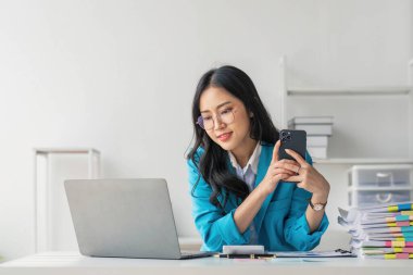 Asyalı Kadın girişimci ofiste işleriyle meşgul. Genç Asyalı kadın masasında bilgisayar başında çalışırken akıllı telefondan ya da cep telefonundan konuşuyor....