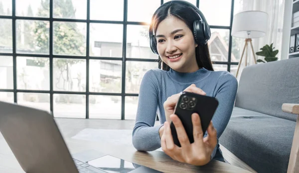 Smiling young asian woman using laptop web camera while wearing headphones and sitting on the rug beside to the sofa at homes..