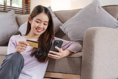 Kredi kartı ve akıllı telefon tutan genç Asyalı kadın tüketici evde oturmuş online bankacılık işlemlerini yapıyor. Sanal alışveriş, güvenli mobil bankacılık kavramı.