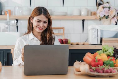 Genç, mutlu, gülümseyen bir kadın, modern mutfakta vegan yemekleri pişirirken dizüstü bilgisayar kullanıyor, gülümseyen hippi kız sağlıklı vitamin salatasını sebzelerle hazırlıyor, sağlıklı yaşam tarzını yiyor...