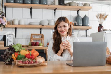 Genç, mutlu, gülümseyen bir kadın, modern mutfakta vegan yemekleri pişirirken dizüstü bilgisayar kullanıyor, gülümseyen hippi kız sağlıklı vitamin salatasını sebzelerle hazırlıyor, sağlıklı yaşam tarzını yiyor...