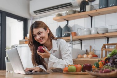 Genç, mutlu, gülümseyen bir kadın, modern mutfakta vegan yemekleri pişirirken dizüstü bilgisayar kullanıyor, gülümseyen hippi kız sağlıklı vitamin salatasını sebzelerle hazırlıyor, sağlıklı yaşam tarzını yiyor...