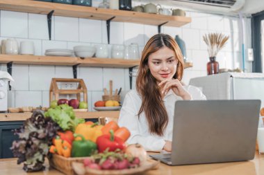 Genç, mutlu, gülümseyen bir kadın, modern mutfakta vegan yemekleri pişirirken dizüstü bilgisayar kullanıyor, gülümseyen hippi kız sağlıklı vitamin salatasını sebzelerle hazırlıyor, sağlıklı yaşam tarzını yiyor...