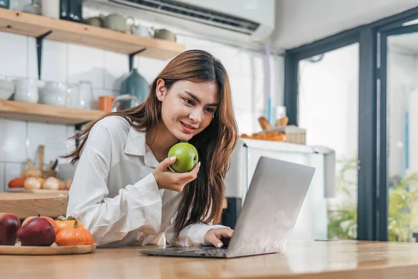 stock image Young happy smiling woman using laptop while cooking vegan food in the modern kitchen, smiling hipster girl preparing healthy vitamin salad with vegetables, eating healthy lifestyle..