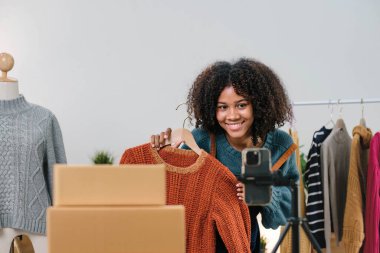 Genç Asyalı kadın moda tasarımcısı cep telefonu siparişi alıyor ve mağazada canlı yayın yapan kıyafetleri gösteriyor. Küçük işletme sahibi, çevrimiçi pazar dağıtımı kavramı...
