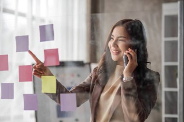 Young smiley attractive, businesswoman using sticky notes in glass wall to writing strategy business plan to development grow to success..