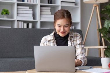 Güzel ve ilham verici Asyalı kadın hafta sonları oturma odasında dizüstü bilgisayarında çalışıyor....