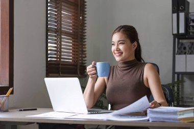 Ofiste rahat bir fincan kahve içerken bilgisayarla çalışan Asyalı mutlu bir iş kadını....
