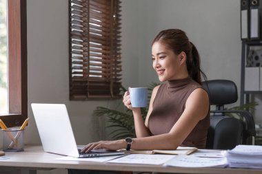 Ofiste rahat bir fincan kahve içerken bilgisayarla çalışan Asyalı mutlu bir iş kadını....