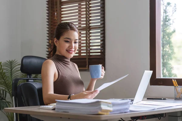 Ofiste rahat bir fincan kahve içerken bilgisayarla çalışan Asyalı mutlu bir iş kadını....