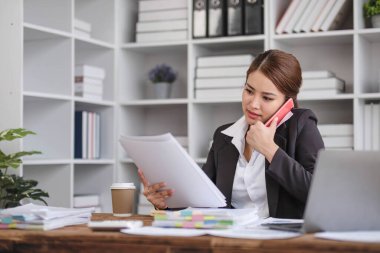 Genç Asyalı iş kadını güzel büyüleyici gülümseyen ve cep telefonuyla konuşan ve ofiste dizüstü bilgisayar kullanan....