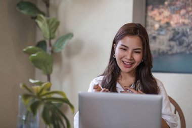 Çekici ve mutlu Asyalı kız kitap okuyor ve oturma odasındaki bir masada dizüstü bilgisayar kullanıyor. Hobi ve boş zaman kavramları..