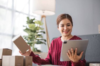Genç bir Asyalı kadının portresi, arka planda paketlerle dolu ofiste oturan bir e-ticaret çalışanı hesap makinesi ve akıllı telefon kullanarak KOBİ iş ekommersi ve