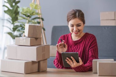 Genç bir Asyalı kadının portresi, arka planda paketlerle dolu ofiste oturan bir e-ticaret çalışanı hesap makinesi ve akıllı telefon kullanarak KOBİ iş ekommersi ve