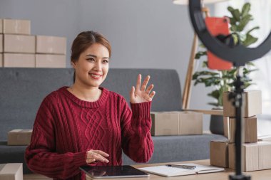 Genç bir Asyalı kadının portresi, arka planda paketlerle dolu ofiste oturan bir e-ticaret çalışanı hesap makinesi ve akıllı telefon kullanarak KOBİ iş ekommersi ve