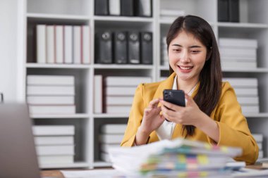 Portre gülüşü güzel Asyalı tasarımcı kadın sarı takım elbiseli ofis masasında telefon bilgisayarı kullanıyor. Küçük işletme çalışanı serbest internet pazarlama e-pazarlama