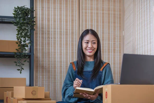 Young beautiful happy asian business woman owner of SME online using laptop receive order from customer with parcel box packaging at her startup home office, online business seller and delivery