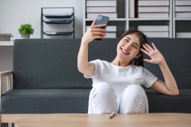 Genç ve mutlu bir Asyalı kadının oturma odasında otururken cep telefonuyla selfie çekmesi....