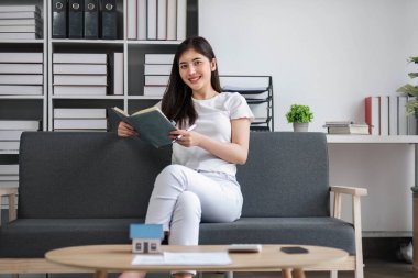 Çekici genç Asyalı kadın en az ve rahat oturma odasında oturup kitap okumanın keyfini çıkarıyor....