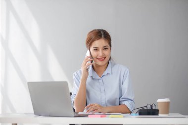 Çekici esmer Asyalı kadın modern ofiste çalışırken cep telefonu kullanıyor, gülümsüyor ve mutlu oluyor....