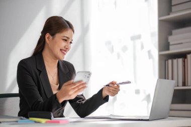 Çekici esmer Asyalı kadın modern ofiste çalışırken cep telefonu kullanıyor, gülümsüyor ve mutlu oluyor....