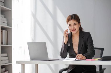 Çekici esmer Asyalı kadın modern ofiste çalışırken cep telefonu kullanıyor, gülümsüyor ve mutlu oluyor....