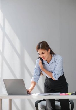 Çekici esmer Asyalı kadın modern ofiste çalışırken cep telefonu kullanıyor, gülümsüyor ve mutlu.
