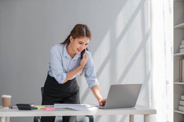 Çekici esmer Asyalı kadın modern ofiste çalışırken cep telefonu kullanıyor, gülümsüyor ve mutlu.